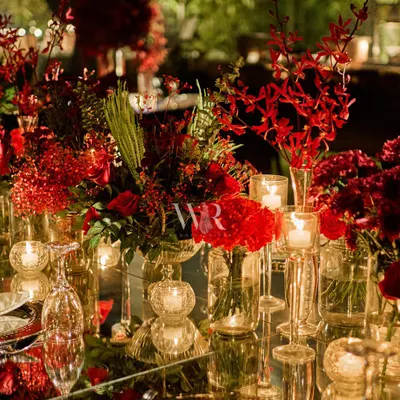 A close-up of a mirrored tablescape, where a runner of red orchids and roses is reflected beautifully. The crystal glassware and silver charger plates add to the layers of luxury and refinement.