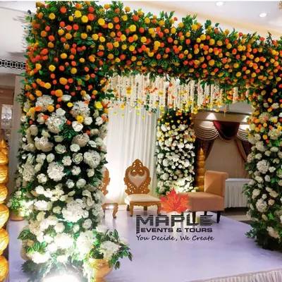 A beautiful mandap setup for a wedding ceremony, featuring pillars wrapped in white and marigold flowers. The traditional seating for the bride and groom is set for the sacred rituals to begin.