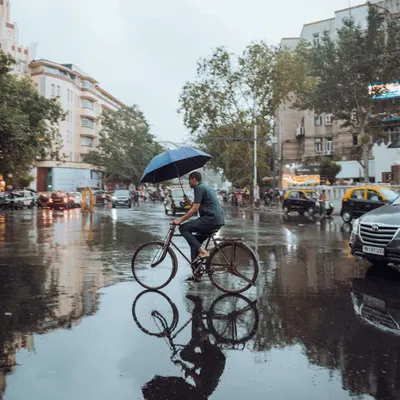A man cycles through a waterlogged Mumbai street during the monsoon, holding an umbrella to balance against the rain. This shot perfectly encapsulates the 'Mumbai grit' and the resilience of its people.