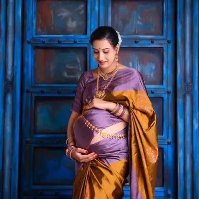 A beautiful solo portrait of the mom-to-be in a traditional purple and gold silk saree. She cradles her bump, radiating grace and serenity against a rustic blue door.