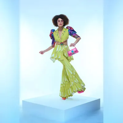 A designer fashion shoot showcasing a vibrant lime green co-ord set. The model's pose is dynamic, highlighting the ruffled details and unique silhouette of the outfit against a clean, ethereal blue-and-white backdrop.