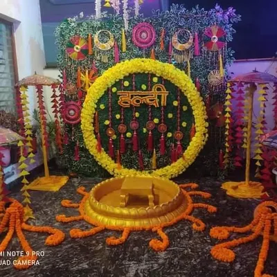 A classic Haldi ceremony stage, featuring a circular backdrop with marigold flowers and the 'Haldi' sign in Hindi. The setup includes a central seating area for the main ceremony.
