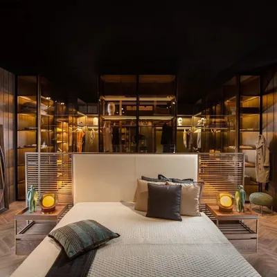 This see-through island wardrobe acts as a functional partition, elegantly dividing the bedroom from the walk-in closet. It is fabricated entirely in glass and metal, allowing for two-way viewing and making the entire space feel larger and more connected.