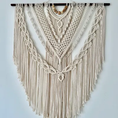 This large, V-shaped macrame wall hanging is made entirely with natural off-white cord and features wooden beads for an earthy touch. The design includes multiple layers of intricate knots, creating amazing texture and a classic bohemian look for any large wall.
