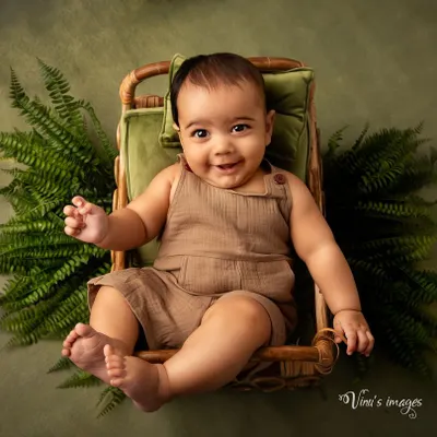 A cheerful smile from a little one. This infant portrait, taken between 2 to 5 months, captures a happy baby sitting in a wicker chair surrounded by green ferns.