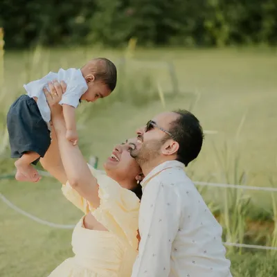 A joyful moment of a mother lifting her baby into the air during an outdoor family photoshoot. The natural light and green surroundings create a fresh and happy scene.