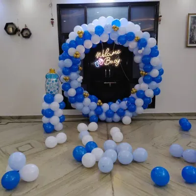 A complete "Welcome Boy" decoration in a living room. This setup includes a circular balloon arch with a neon sign, loose balloons on the floor, and a balloon pillar with a baby bottle foil balloon.