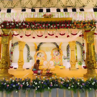 A magnificent wedding mandap with ornate golden pillars and a backdrop featuring deities. The stage is heavily decorated with flowers, creating a divine setting for the marriage ceremony.