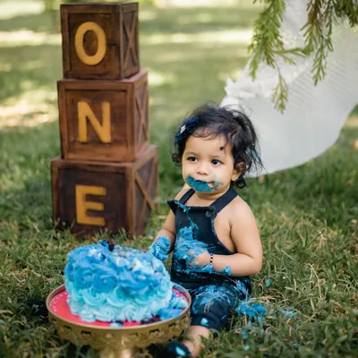 A one-year-old gets delightfully messy with a blue birthday cake during an outdoor cake smash session. The rustic "ONE" blocks and teepee in the background create a perfect boho-chic setup.