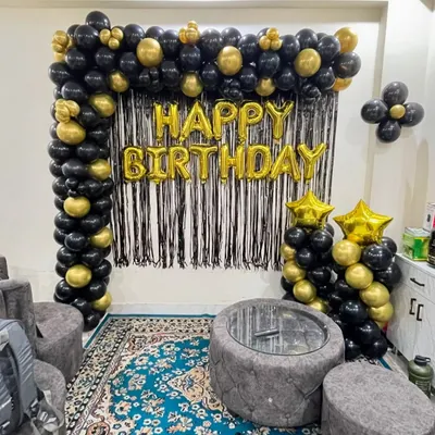A classic and elegant birthday decoration for a living room. This setup features a black and gold balloon arch over a black fringe backdrop, with "Happy Birthday" foil letters.