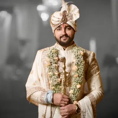 A groom ready for his ceremony, wearing a classic ivory sherwani. The outfit is detailed with fine embroidery and paired with a traditional safa and a ceremonial sword, completing the royal look.