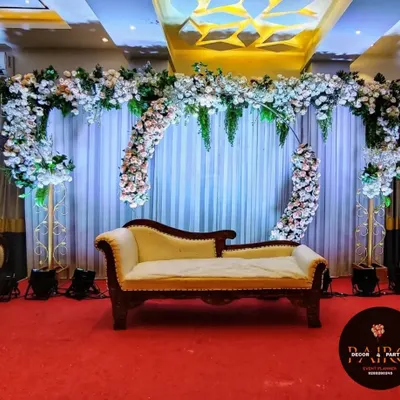 This elegant stage decoration for a ring ceremony features a beautiful arch of white and blush pink flowers. A classic chaise lounge provides sophisticated seating for the couple against a simple white curtain backdrop.