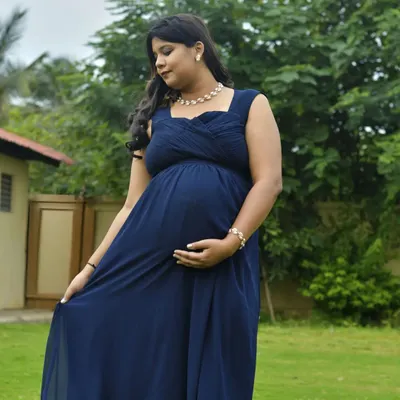 A serene outdoor portrait in a beautiful garden. The mother-to-be, dressed in a flowing navy gown, looks down at her bump in a moment of quiet reflection, surrounded by nature.
