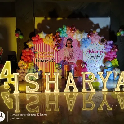 This 'Candyland' themed birthday party features large, light-up marquee letters spelling 'AISHARYA'. The backdrop includes a colorful organic balloon garland with donut and candy accents to bring the sweet theme to life.