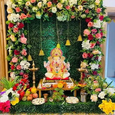 A complete Ganpati decoration featuring a dense floral frame of pink, white, and red roses around an artificial green wall. The hanging bells and traditional lamp create a perfect setting for pooja.