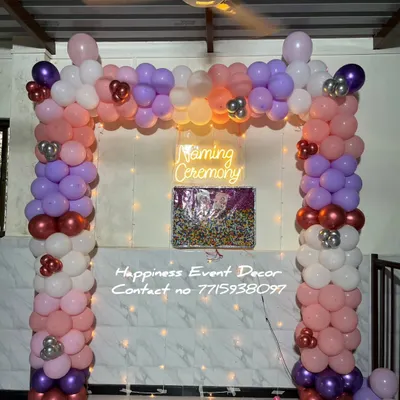 This is a popular decoration for a naming ceremony at home. It features a full balloon arch in multiple colors with a 'Naming Ceremony' neon sign and a name reveal board against a simple tiled wall.