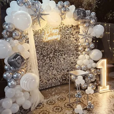 A glamorous first-anniversary party with a silver shimmer wall, a "Happy Anniversary" neon sign, and a silver and white balloon arch. The light-up number '1' marks the special milestone.