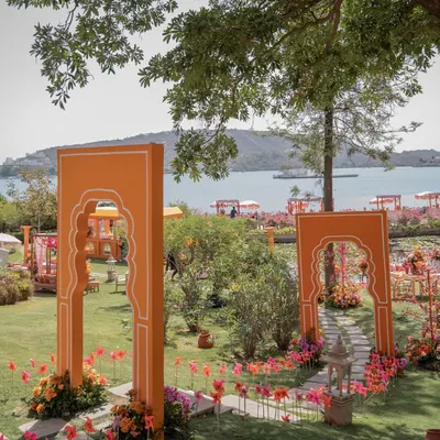 The entrance to the 'Pyaar ka Pind' Haldi, with vibrant orange arches framing a stunning view of Lake Pichola in Udaipur. This sets the stage for a colorful, destination celebration.