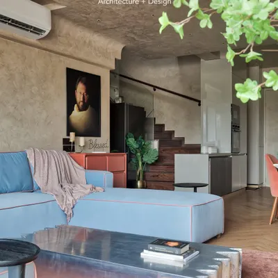 The living room of the Red Fort Project uses a calm, soft color palette to create a tranquil atmosphere. The light blue L-shaped sofa with red piping adds a subtle pop of color against the textured beige walls.