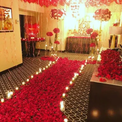 A luxurious room decoration for a proposal, featuring a dense pathway of red rose petals, candlelights, and red foil balloons creating a deeply romantic ambiance.