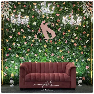 This reception stage is a perfect blend of nature and glamour. A lush green fern wall is adorned with pastel flowers, sparkling chandeliers, and the couple's initials, with a luxurious velvet sofa taking center stage.
