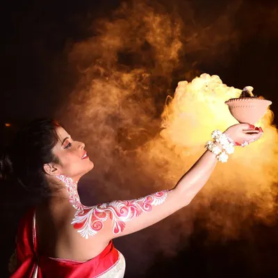 A creative shot for Durga Puja, where the model holds a dhunuchi (incense burner). The traditional Alpona body art on her arm is prominently displayed, glowing against the smoky background.