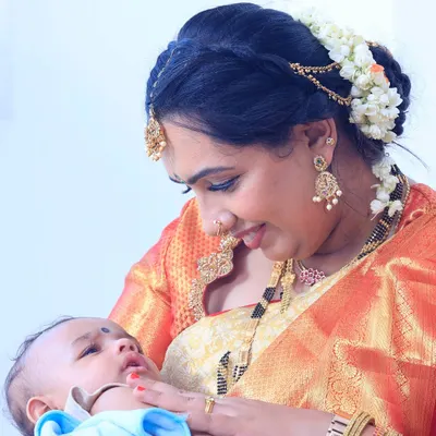 A precious moment between mother and child at her baby boy's naming ceremony. Her makeup is soft and radiant, with a beautiful floral bun, perfect for this heartfelt occasion.