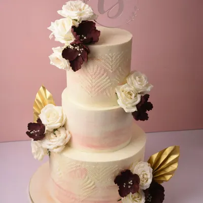 A stunning three-tier wedding cake with a soft peach ombré effect and delicate leaf-stamped texture. It's adorned with white and deep burgundy sugar flowers and gold leaf accents for a touch of modern romance.