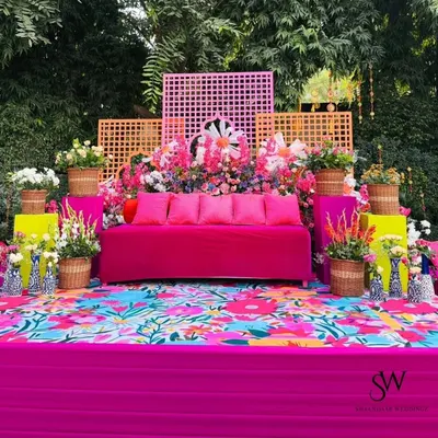 A vibrant and colorful Mehendi or Haldi seating area. The bright pink sofa, floral printed flooring, and abundant flowers in wicker baskets create a festive and cheerful atmosphere.