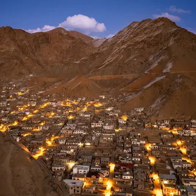 An aerial view of a town in Ladakh as dusk settles, with lights beginning to glow in the valley. This shot captures the quiet beauty of human settlement nestled amongst the towering, barren mountains of the Himalayas.