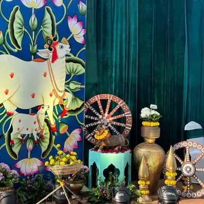 A beautiful setup for a Radha Krishna themed event, featuring a custom backdrop with Pichwai art of a cow and calf. The decor is complemented by brass pots, painted wheels, and marigold flowers.