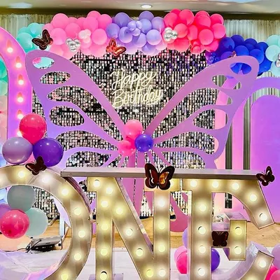A magical first birthday setup combining a butterfly theme with large "ONE" marquee letters. The decor features a silver shimmer wall, colorful balloon arrangements in teal, pink, and purple, and detailed butterfly cutouts.