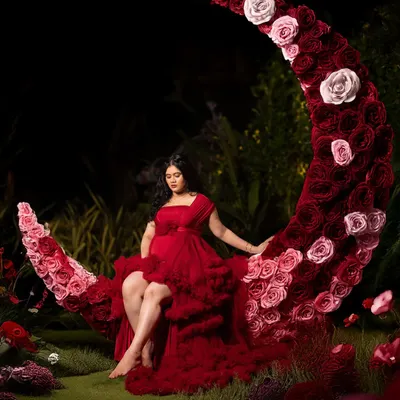 A portrait of pure fantasy. The mom-to-be sits upon a floral crescent moon, dressed in a bold red gown, creating a magical and artistic image in our garden studio after sunset.