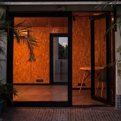 The entrance to a small workspace at dusk. The warm interior light glows through the full-height glass door, creating an inviting threshold between inside and out.