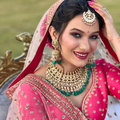 A picture-perfect bride with a dazzling smile. Her makeup features smoky eyes and a bold berry lip, beautifully contrasting with her bright pink lehenga and emerald green jewellery.