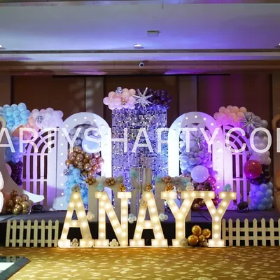 This adorable teddy bear stage is perfect for a first birthday. The setup features two giant teddy bear props, a sparkling sequin backdrop, and a white picket fence in front of the illuminated name 'ANAYY', creating a cute and cuddly atmosphere.