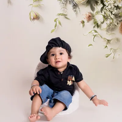A simple and elegant studio portrait. The baby is dressed in a stylish dark shirt and cap, set against a clean white background with a delicate floral arrangement overhead.
