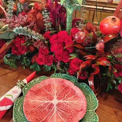 A close-up of a place setting from our "Scarlet House" theme. A vibrant, custom-printed plate sits atop a green charger, complemented by a bold red napkin and a backdrop of lush, textured red florals.