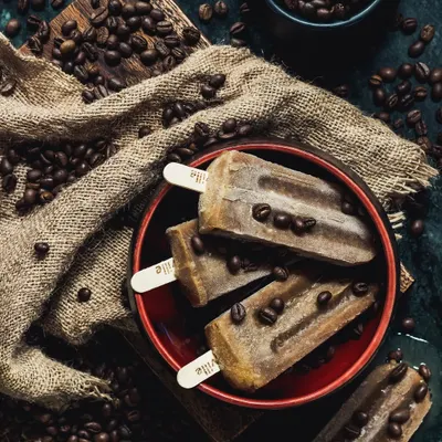 Coffee-flavored popsicles styled in a dark, moody setting with scattered coffee beans. This conceptual shot is designed to be artistic and attention-grabbing.