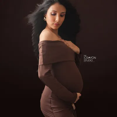 A fine art portrait against a rich, dark backdrop. The soft light highlights the curve of her bump and the gentle movement of her hair, creating a poetic and graceful image.