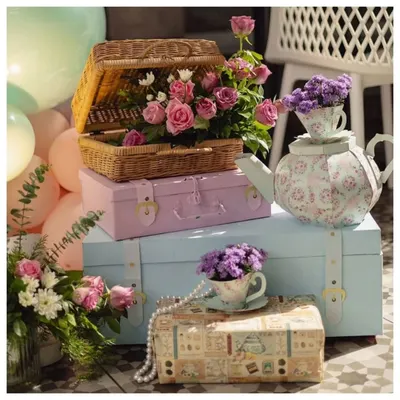 A beautiful collection of props for a vintage tea party theme. The display includes pastel-colored storage trunks, a wicker picnic basket filled with roses, and a floral teapot centerpiece.