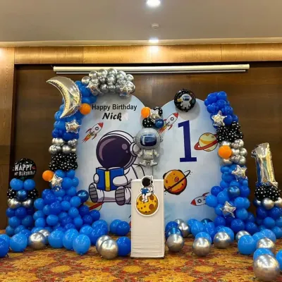 A complete astronaut-themed stage for Nick's first birthday. This setup includes a large custom backdrop with space graphics, a full balloon garland with silver and orange accents, and a themed cake stand.