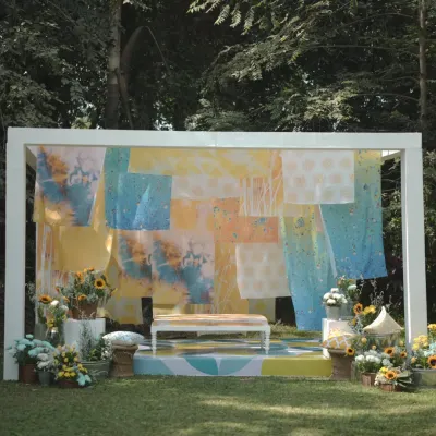 A full view of the unique "Dhobi Ghat" themed Haldi setup. The backdrop of colorful tie-dyed fabrics, combined with a patterned stage floor and sunflower arrangements, creates a vibrant and artistic atmosphere.