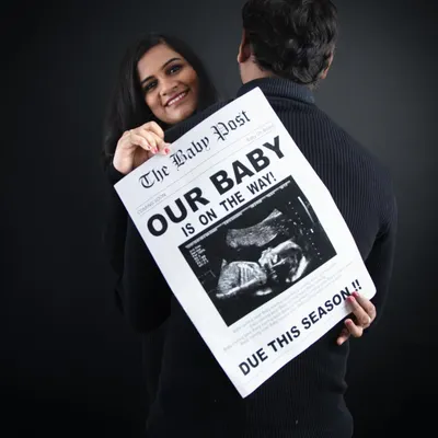 A playful and creative announcement. The mom-to-be peeks over her partner's shoulder while he holds a custom newspaper prop announcing their baby is on the way.