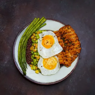 A high-protein meal with grilled chicken, two sunny-side-up eggs, quinoa, and fresh green beans, shot with dramatic lighting.