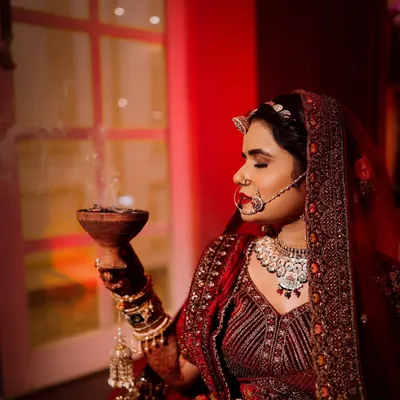 A serene bridal portrait showing the bride in her red lehenga, holding a traditional bowl with incense smoke. The warm, red lighting creates a spiritual and deeply cultural atmosphere for this wedding day photo.