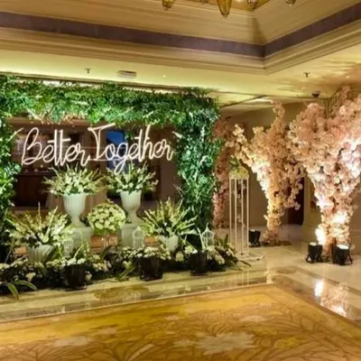 A beautiful photo corner for an engagement party with a 'Better Together' neon sign. The backdrop of lush greenery and cherry blossom trees creates a romantic and whimsical setting.