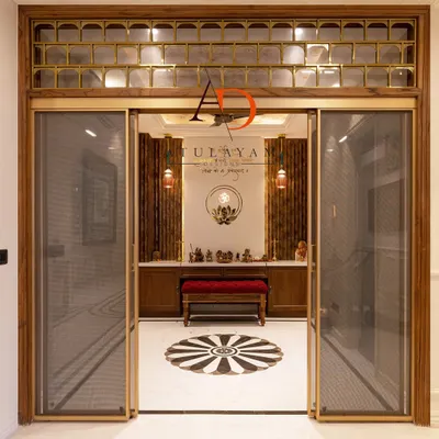 This grand pooja room entrance features sliding glass doors with a fine mesh and a transom with brass and glass detailing. The design creates an inviting yet separate sacred space, with a view of the beautiful marble floor inlay inside.