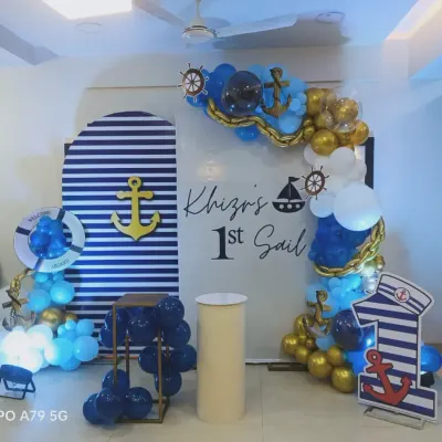 A nautical-themed decoration for "Khiza's 1st Sail". This setup features a striped backdrop, an anchor cutout, a ship's wheel, and a blue and gold balloon arch, perfect for a little sailor's party.