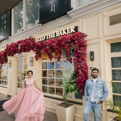 A modern fairytale scene in front of a beautiful bakery. The contrast between her flowing pink gown and his casual denim creates a stylish, contemporary pre-wedding portrait.